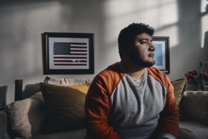 Man sitting in a darkened room, a small framed American flag on the wall behind